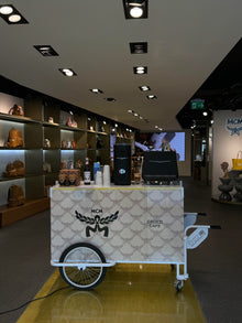 MCM branded coffee cart in a store setting with shelves and a screen in the background.