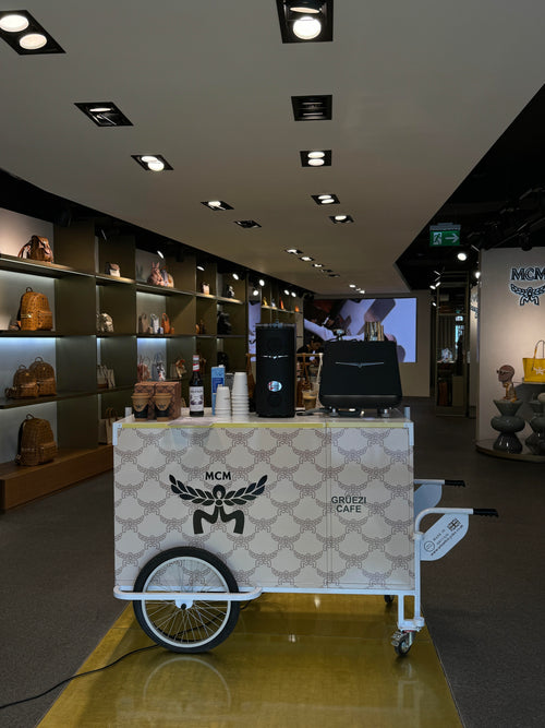 MCM branded coffee cart in a store setting with shelves and a screen in the background.